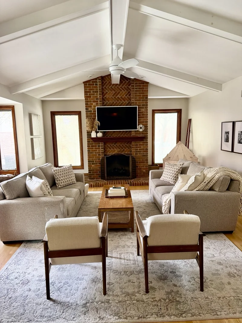 Rochester Hills living room with a vaulted white ceiling, brick fireplace, neutral sofas, and wood framed chairs